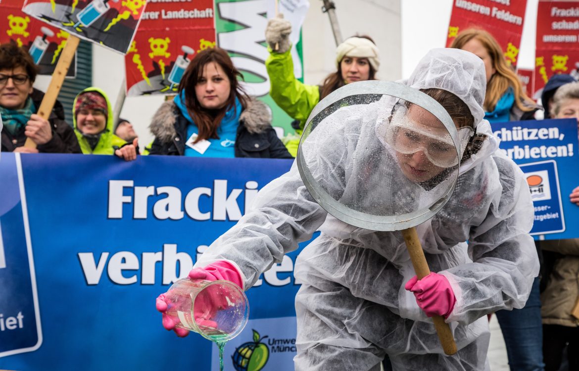 Demonstrantin bei Demonstration in Laborkleidung und großer Lupe untersucht Fracking