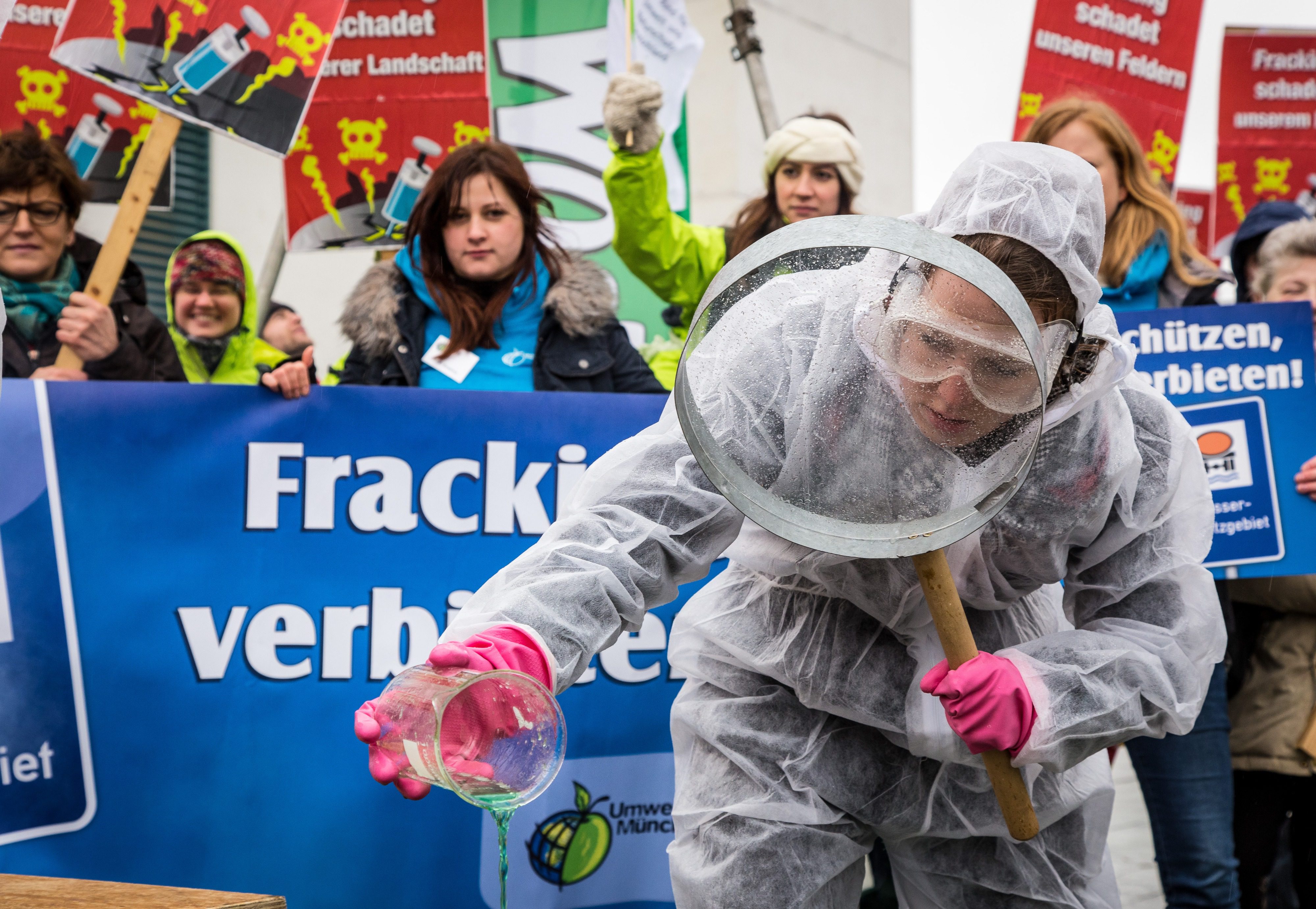 Demonstrantin bei Demonstration in Laborkleidung und großer Lupe untersucht Fracking