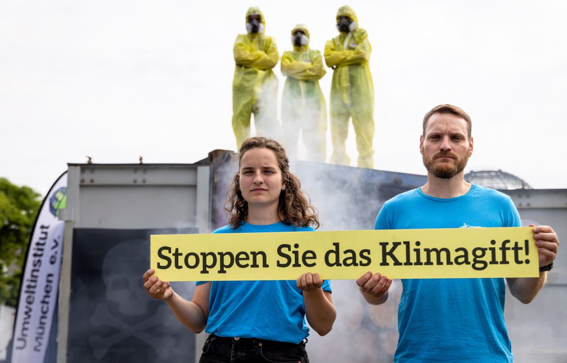 Referent:innen des Umweltinstitut vor rauchendem Container mit Schild: 