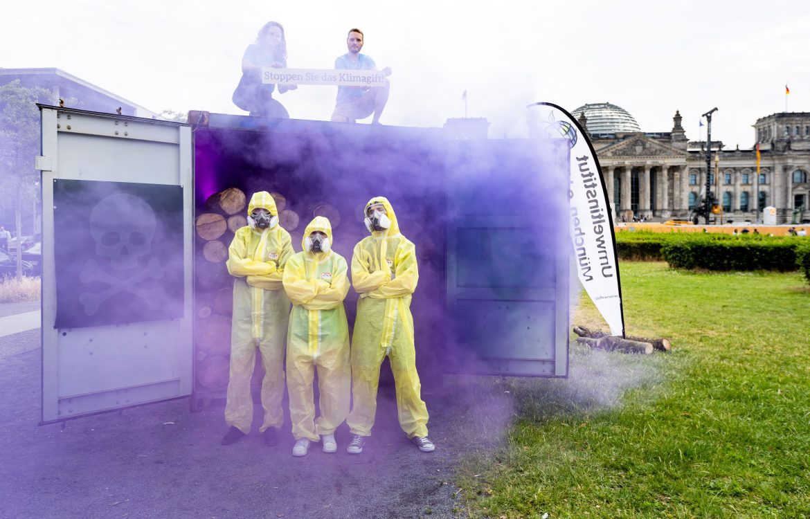 Drei Menschen in gelben Schutzanzügen stehen vor einem Container, aus dem lila Gas strömt. Auf dem Dach des Containers halten zwei Menschen ein Schild mit der Aufschrift 