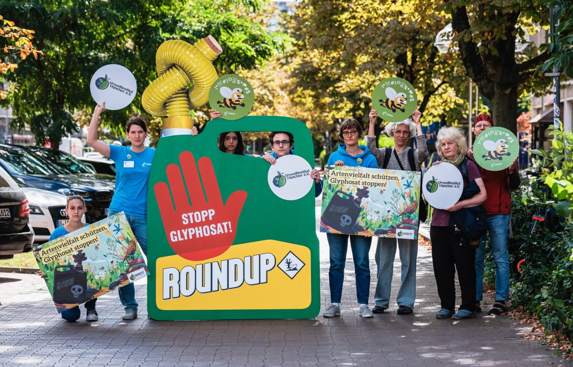 Das Team vom Umweltinstitut München bei der Übergabe-Aktion gegen Glyphosat in Berlin 2023 vor dem Landwirtschaftsministerium mit Protestschildern und einem übergroßen Roundup Kanister mit zugeknotetem Schauch