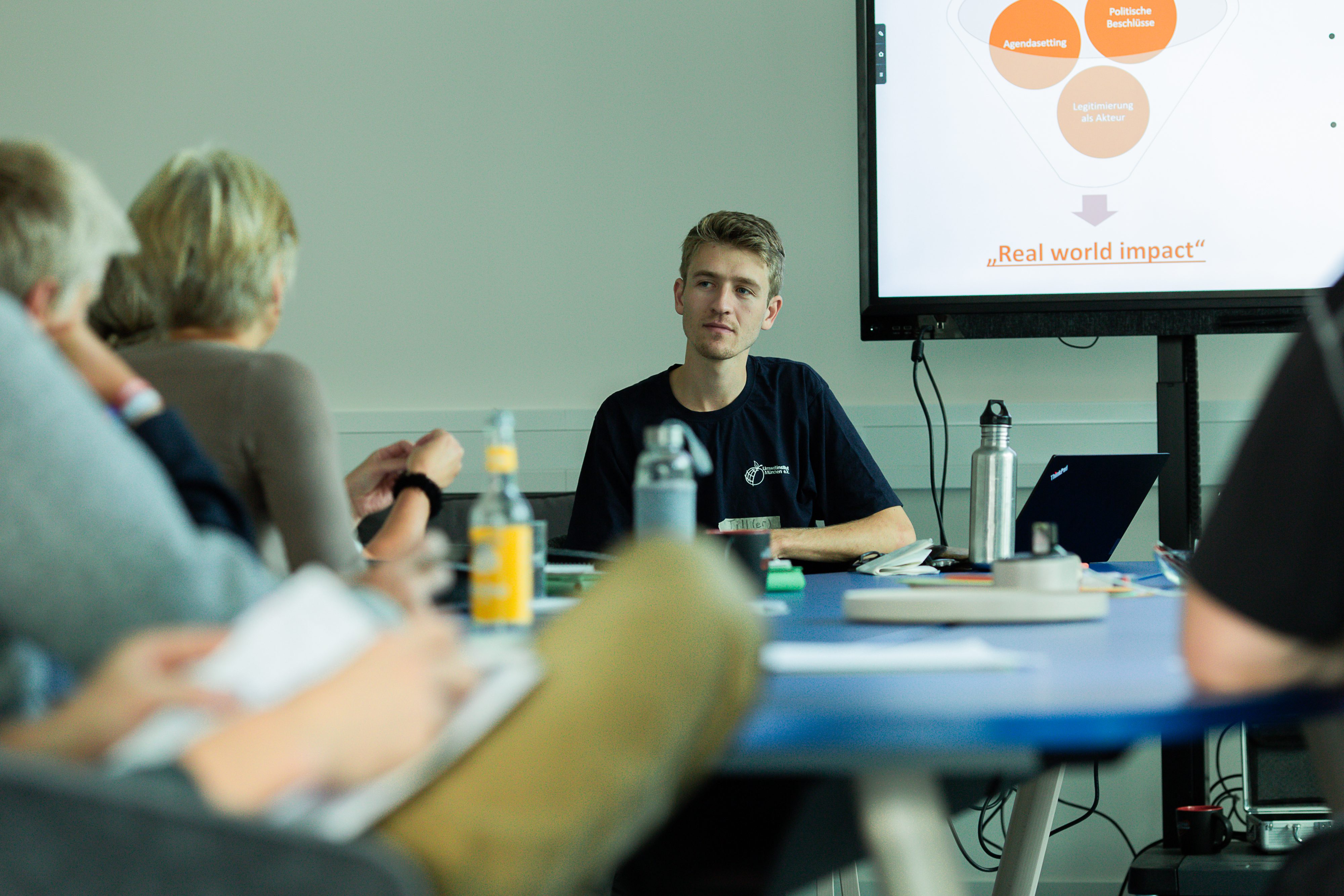 Referent des Umweltinstitut sitzt an einem Tisch mit anderen Beteiligten. Hinter ihm ist ein Bildschirm, auf dem eine Präsentation zu sehen ist.