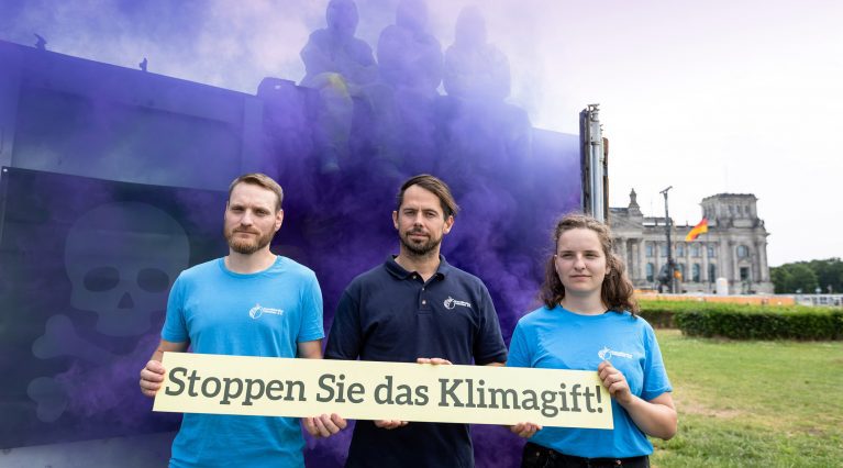 Violetter Rauch strömt aus einem Seecontainer mitten in Berlin, im Vordergrund halten Protestierende ein Schild mit der Aufschrift 