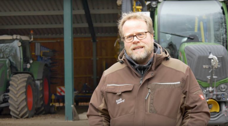 Der Ökolandwirt Reiner Bonhorst (mittelalt, blonde kurze Haare und kurzer Bart, Brille mit braunem Rahmen) steht in einer braunen Jacke vor einer Halle mit Traktoren.