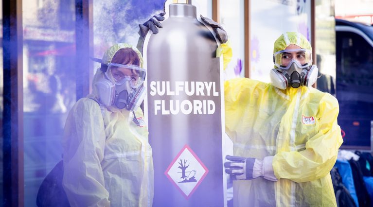 Protest gegen Sulfurylfluorid: Menschen in Schutzmontour halten riesige lila rauchende Gasflasche