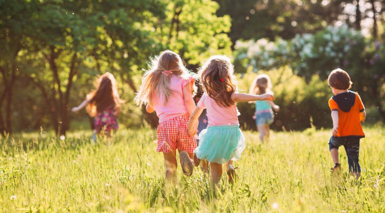 Kinder spielen an einem sonnigen Tag auf einer grünen Wiese, umgeben von Bäumen.