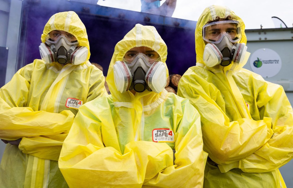 Drei Personen in gelben Schutzanzügen und mit Atemmasken stehen vor einem Container, aus dem lila Nebel entweicht.