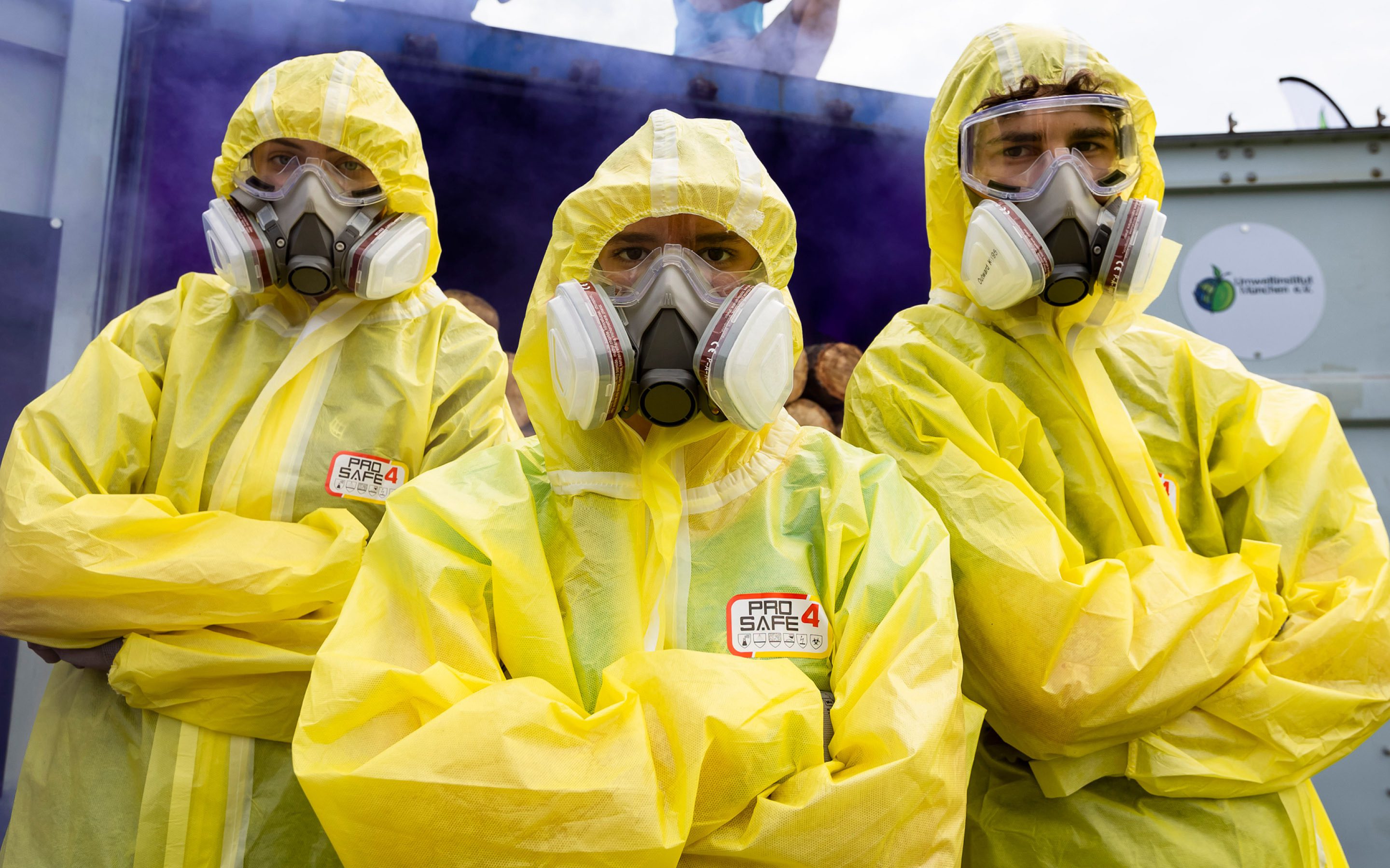 Drei Personen in gelben Schutzanzügen und mit Atemmasken stehen vor einem Container, aus dem lila Nebel entweicht.