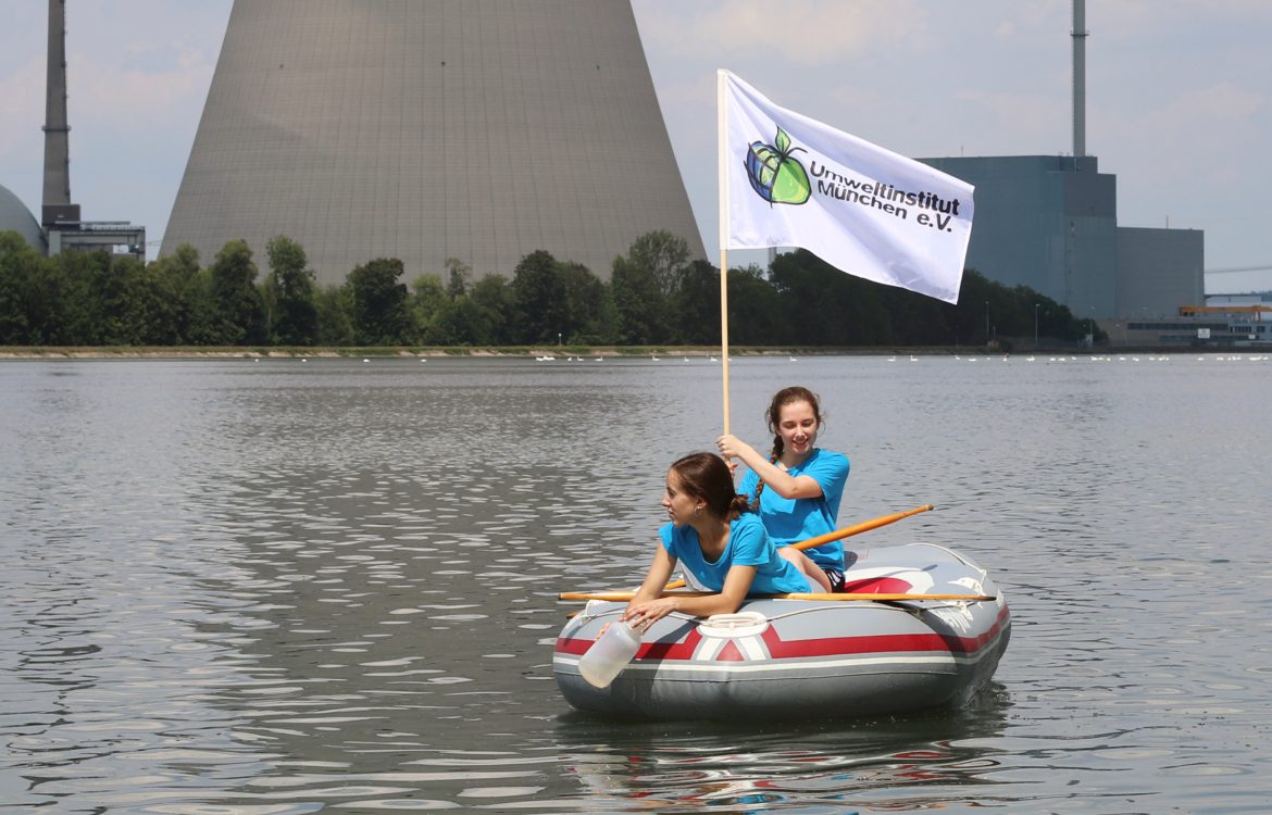 Zwei junge Menschen sitzen in einem Schlauchboot in einem Fluss vor einem Kernkraftwerk.