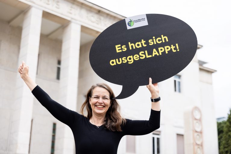 aktuelle meldung_slapp-gesetzesentwurf_c-JörgFarys Frau hält Sprechblasenschild mit Aufdruck ‚Es hat sich ausgeSLAPPt!‘ vor einem hellen Gebäude hoch.