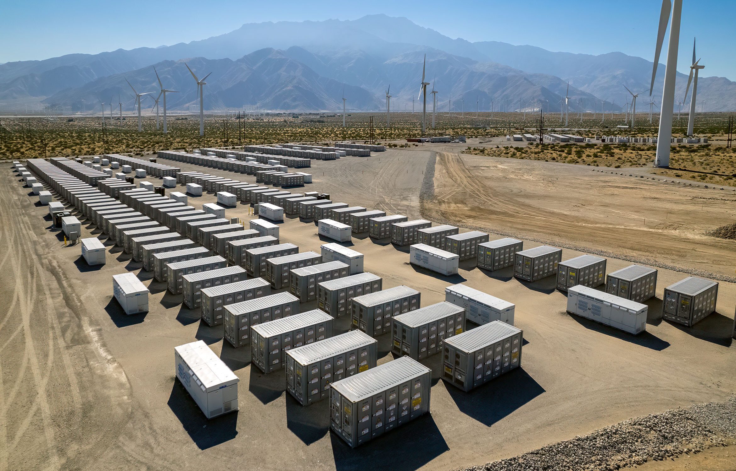 Große Anlage mit Reihen aus grauen Container-Batteriespeichern in einer Wüstenlandschaft, im Hintergrund zahlreiche Windräder und Gebirgskette unter blauem Himmel.