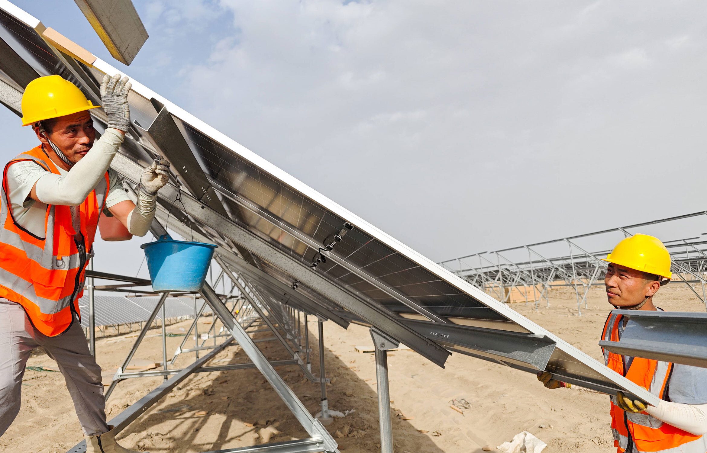Zwei Bauarbeiter in orangenen Warnwesten und gelben Helmen installieren ein Solarmodul auf einer Metallkonstruktion in einer trockenen, sandigen Landschaft.