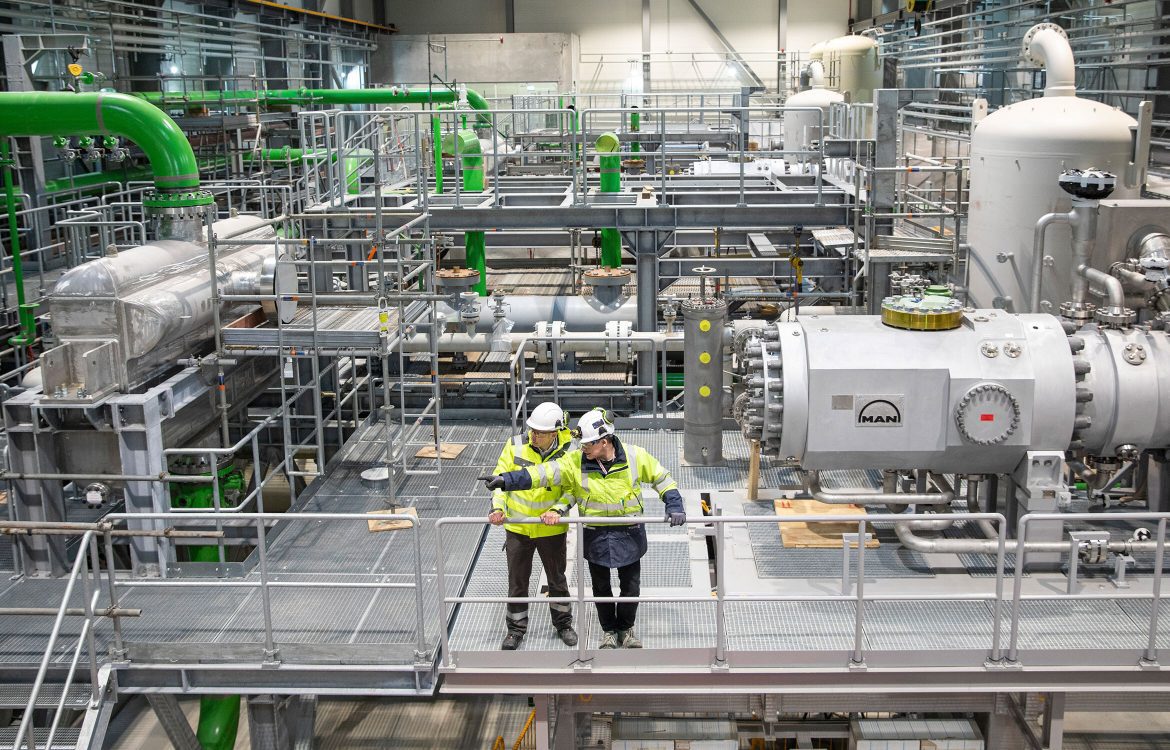 Zwei Personen in Sicherheitskleidung und Helmen stehen in einer modernen Industriehalle mit großen Maschinen, Rohren und Leitungen, teils in Grün gestrichen.