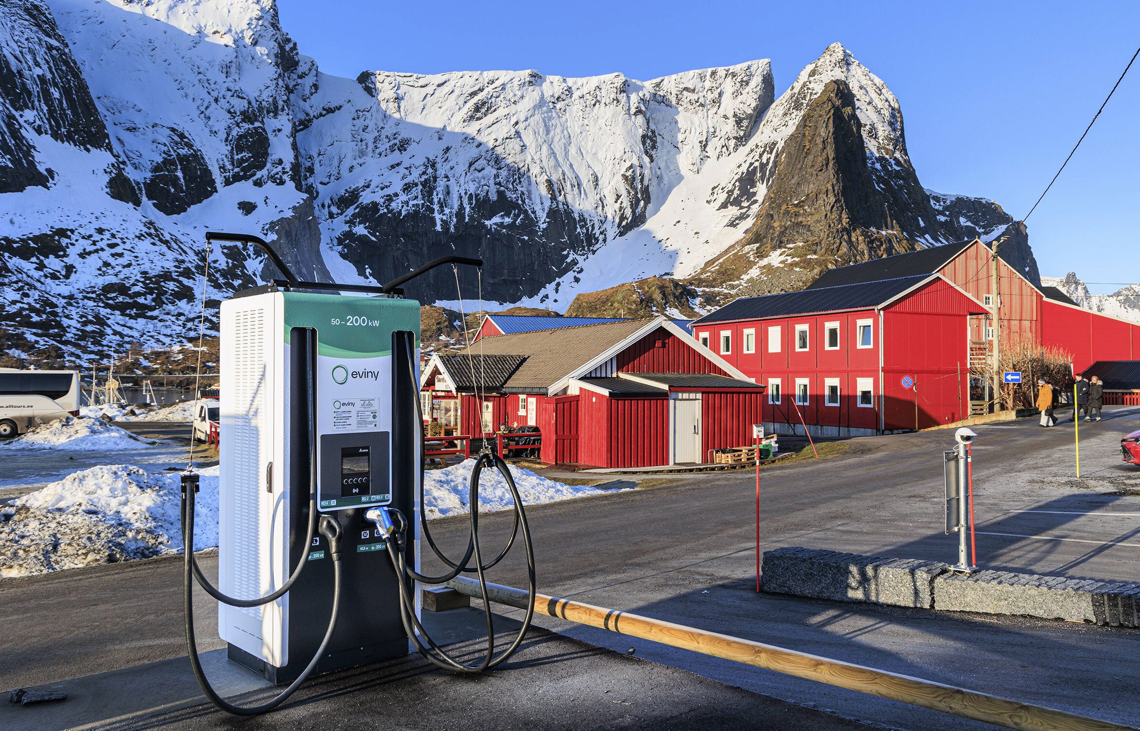 Moderne Elektroauto-Schnellladestation in einem verschneiten Dorf mit roten Holzhäusern, dahinter steile schneebedeckte Felsen unter blauem Himmel.