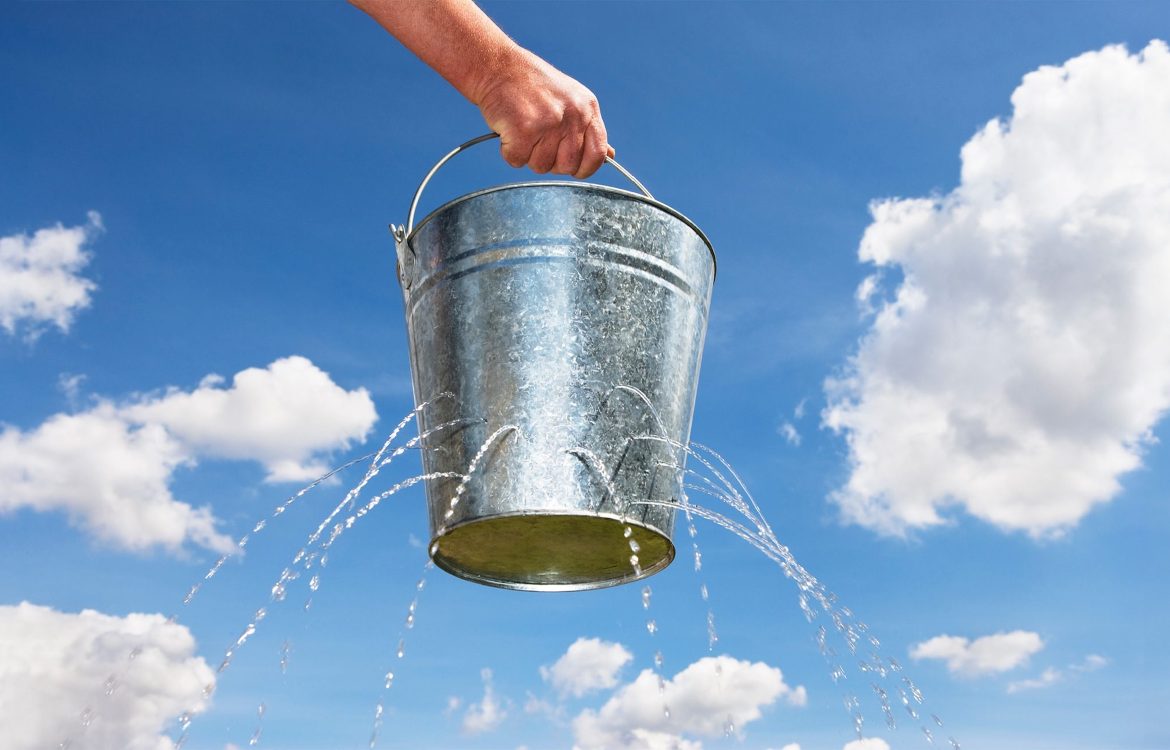 Metalleimer mit Löchern, aus dem Wasser herausläuft, vor blauem Himmel mit Wolken.