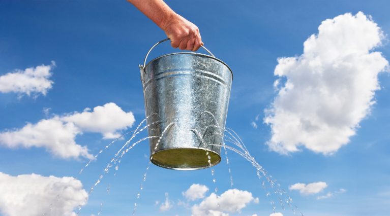 Metalleimer mit Löchern, aus dem Wasser herausläuft, vor blauem Himmel mit Wolken.