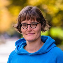 Das Porträt zeigt Sophia Guttenberger, Landwirtschaftsreferentin am Umweltinstitut München. Sie steht im Freien vor einem unscharfen Hintergrund mit herbstlich gefärbten Bäumen. Sie trägt einen blauen Kapuzenpullover und eine runde Brille und blickt ruhig in die Kamera.