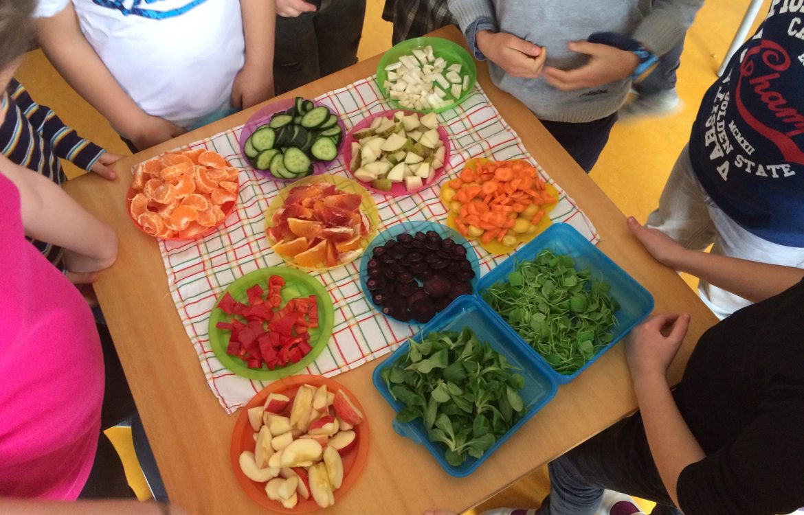 Auf einem Tisch stehen viele Teller mit verschiedenen Obst und Gemüsesorten. Um den Tisch stehen Kinder.