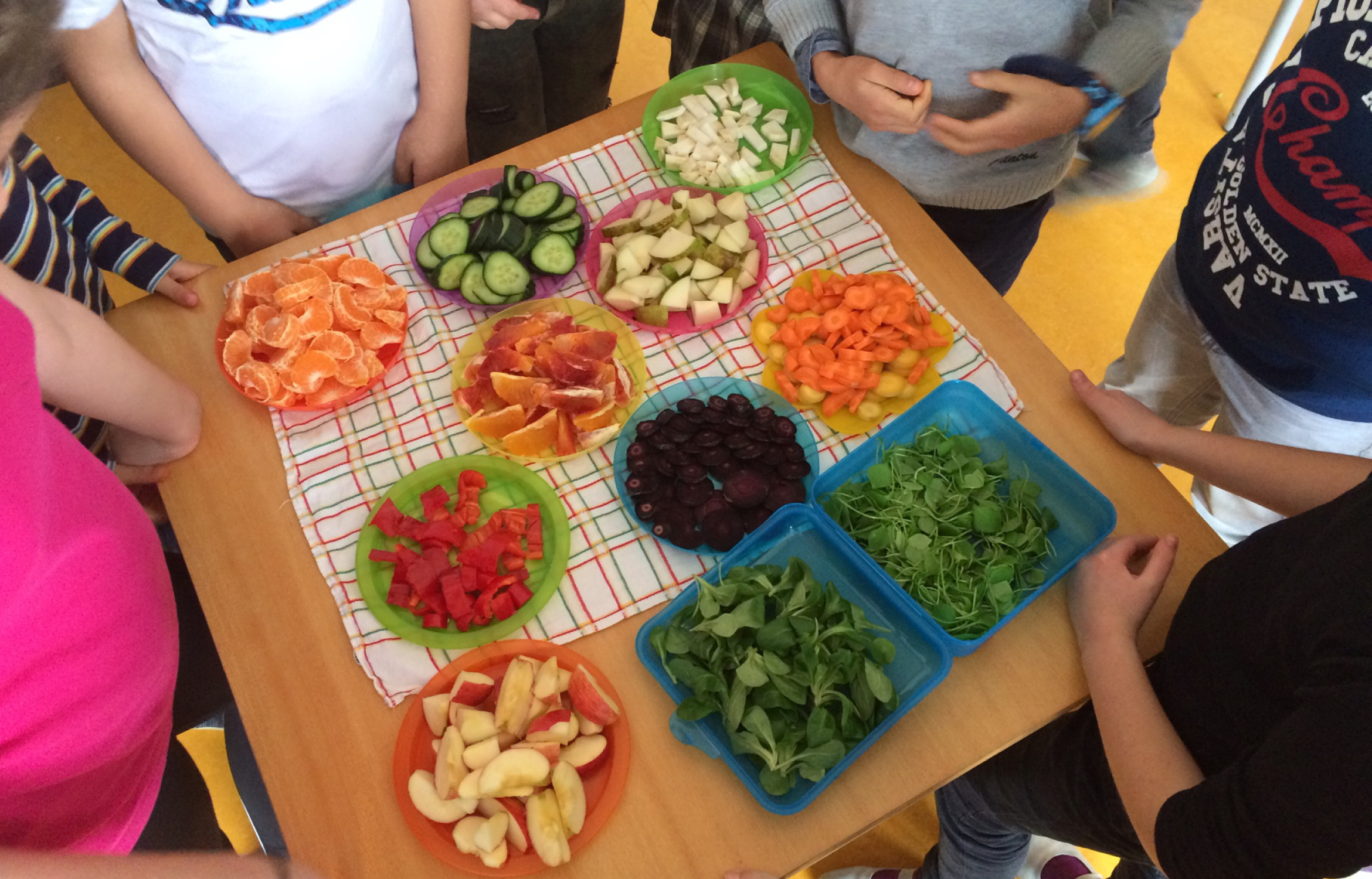 Auf einem Tisch stehen viele Teller mit verschiedenen Obst und Gemüsesorten. Um den Tisch stehen Kinder.