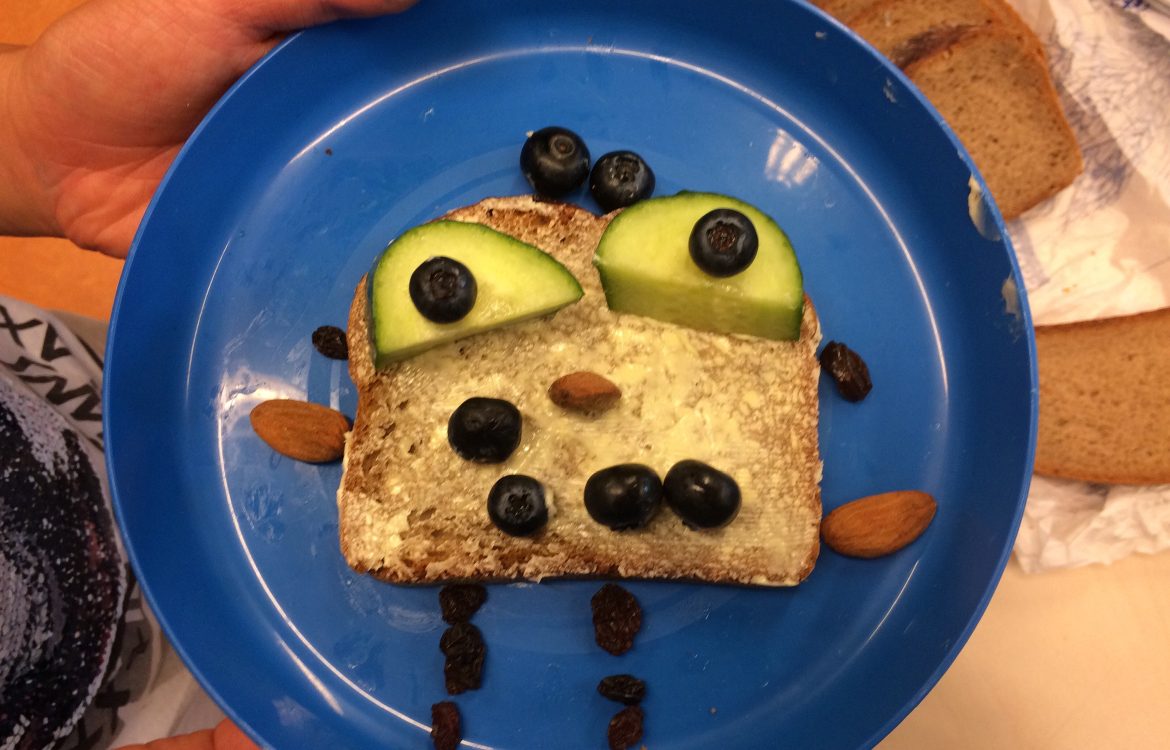 Auf einem Teller liegt eine Scheibe Brot, auf der mit Gemüse und Obst ein lustiges Gesicht gelegt wurde.